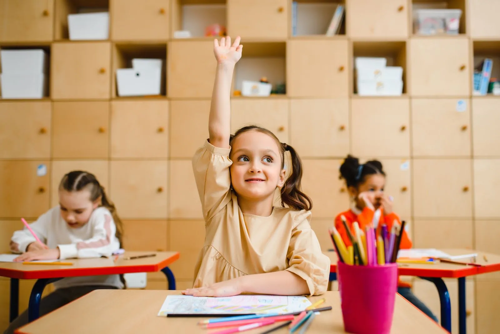 A young girl raising her hand at her desk in a warm, calm classroom